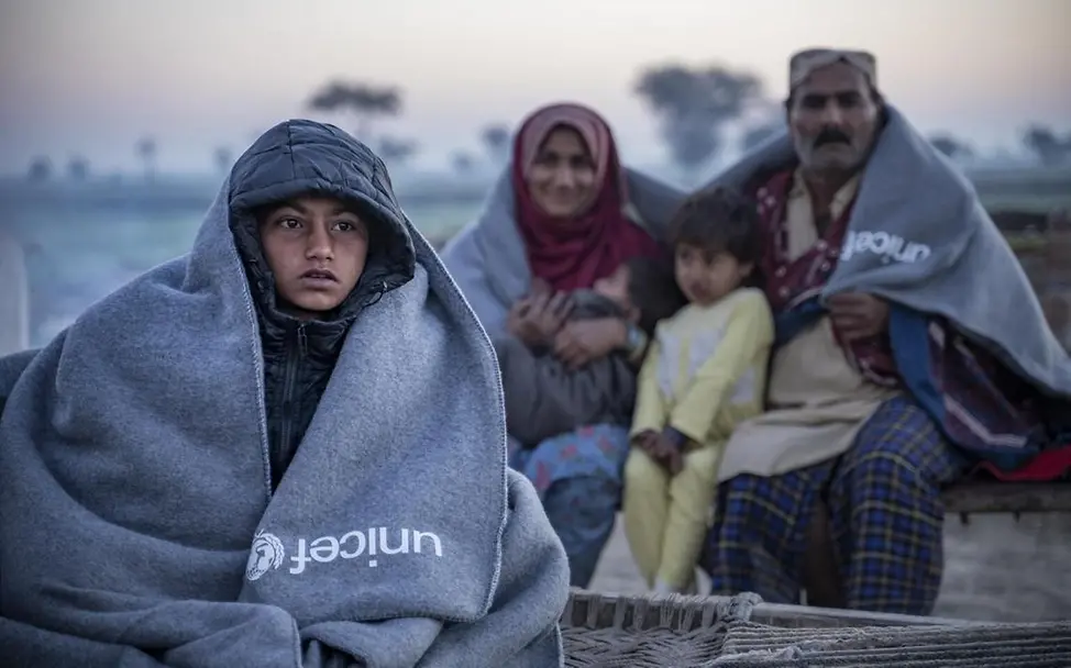 In dem Distrikt Dadu sitzt eine Familie in Decken gehüllt vor ihrem Zuhause. Sie haben von UNICEF ein Paket mit Winterartikeln erhalten. | © UNICEF/Butt Familie sitzt in Decken gehüllt vor ihrem Zuhause.