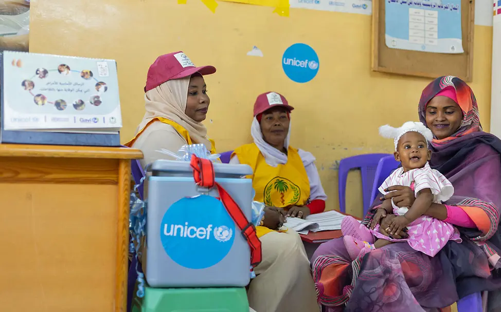 Ein Baby im Sudan wurde gegen Malaria geimpft, um Schutz vor der Infektionskrankheit zu haben | © UNICEF/Elfatih Eine Mutter sitzt mit ihrem Kind vor einer Kiste von UNICEF mit Impfstoff.