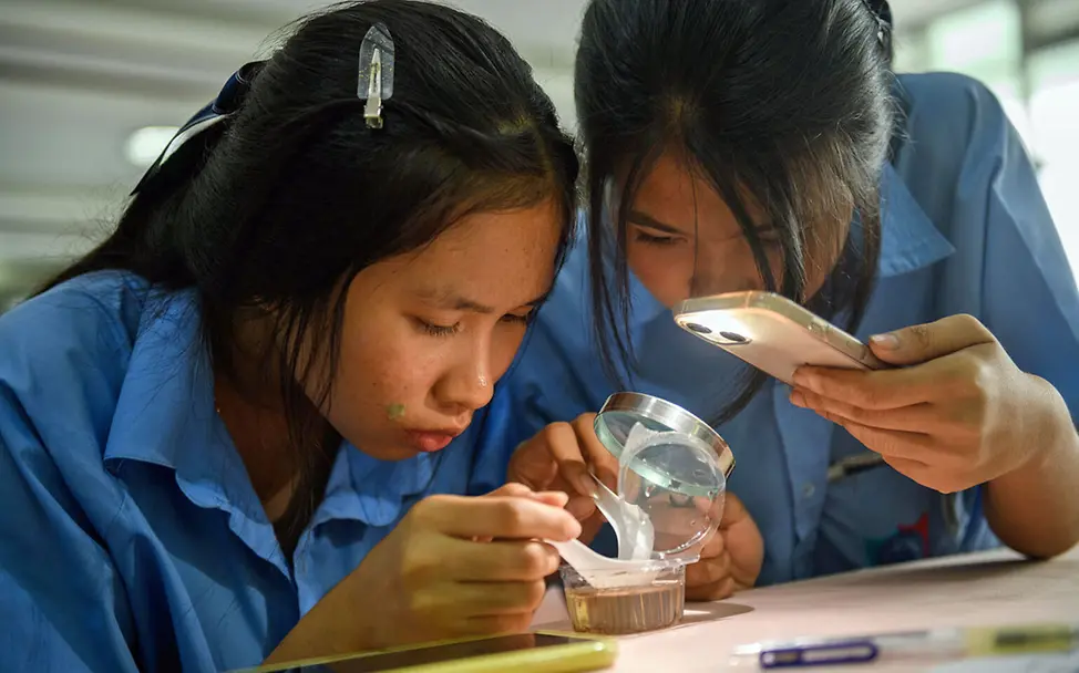 Zwei Schülerinnen in Thailand untersuchen Mückenlarven unter dem Mikroskop | © UNICEF/Roisri Malaria Kinder schützen: Zwei Schülerinnen in Thailand untersuchen mit einer Lupe und einer Handy-Taschenlampe Mückenlarven.