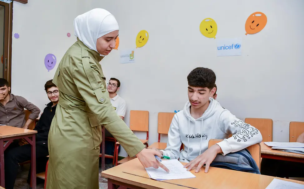 Bilal hat in den Selbstlernzentren auch neue Freunde gefunden. | © UNICEF/ Shahan Der 17-jährige Bilal sitzt in der Schule und seine Lehrerin erklärt ihm etwas zu der Aufgabe auf seinem Tisch.