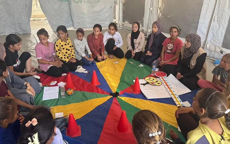 Kinder nehmen an einer von UNICEF unterstützten Aktivität zur psychischen Gesundheit teil. | © UNICEF/Eleyan Kinder und junge Erwachsene sitzen im Kreis auf einer bunten Decke.